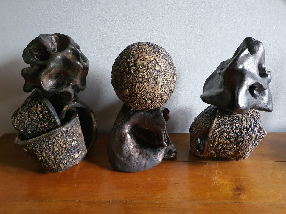 Trzy ciemne rzeźby ceramiczne o bogatej fakturze ustawione obok siebie na drewnianym blacie